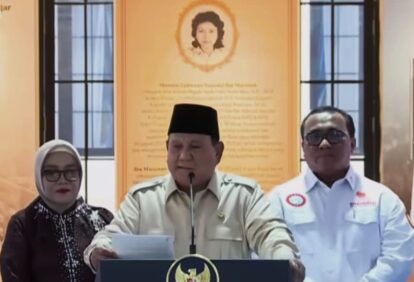 Three people at a podium during a press conference; center man speaks into a microphone, flanked by two colleagues against an orange wall with a portrait.