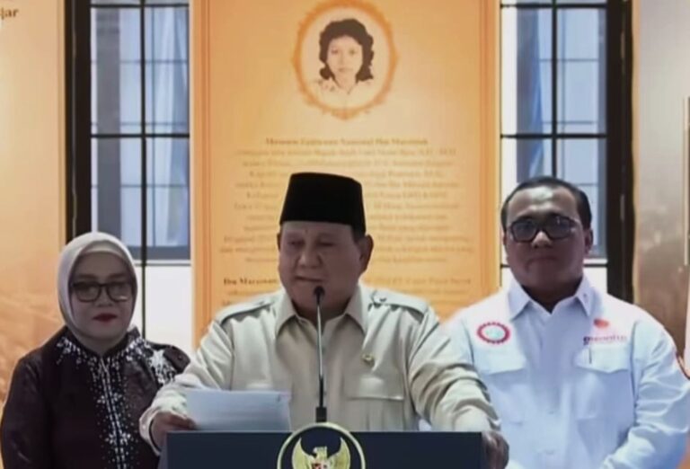Three people at a podium during a press conference; center man speaks into a microphone, flanked by two colleagues against an orange wall with a portrait.
