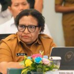 Woman in brown uniform and black glasses seated at a conference table name badge reads ribka haluk with flowers and a laptop nearby