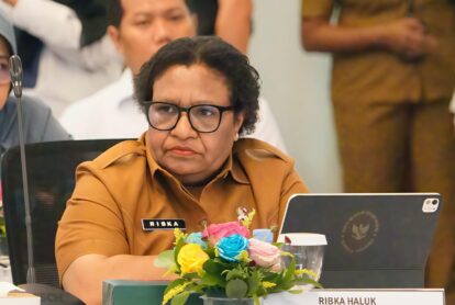 Woman in brown uniform and black glasses seated at a conference table name badge reads ribka haluk with flowers and a laptop nearby