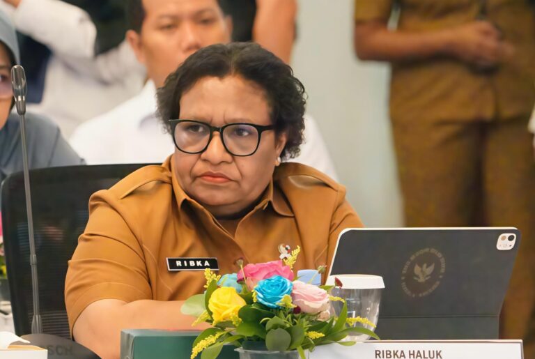 Woman in brown uniform and black glasses seated at a conference table, name badge reads Ribka Haluk with flowers and a laptop nearby.
