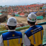 Two construction workers in white safety helmets and blue yellow vests overlook a busy site marked by orange steel framework and piles of materials