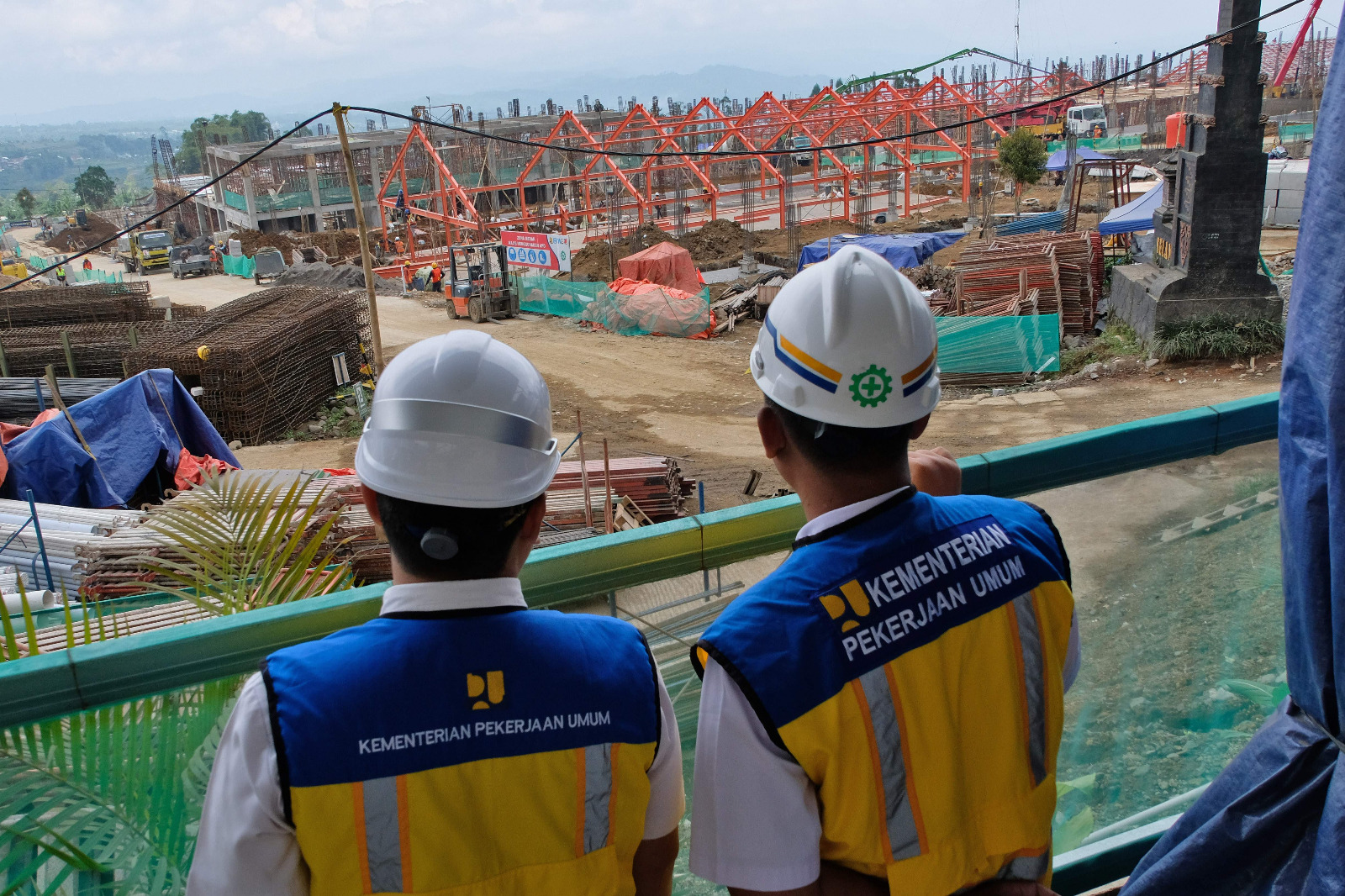 Two construction workers in white safety helmets and blue yellow vests overlook a busy site marked by orange steel framework and piles of materials