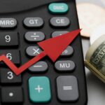 Calculator close up with a red upward arrow and a roll of dollar bills symbolizing financial growth or rising profitsexpenses
