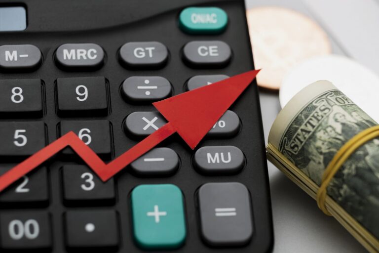 Calculator close-up with a red upward arrow and a roll of dollar bills, symbolizing financial growth or rising profits/expenses