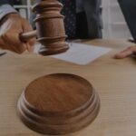 Judges gavel about to strike a wooden sound block on a desk with a hand resting nearby and papers in the background
