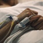Patients arm with iv line connected to a catheter and tape resting on a hospital bed with hands clasped