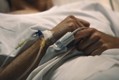 Patients arm with iv line connected to a catheter and tape resting on a hospital bed with hands clasped