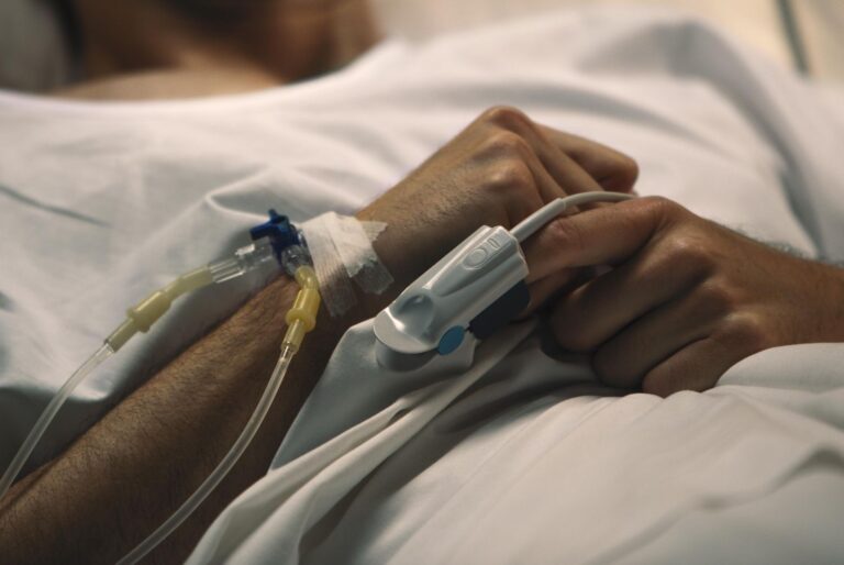 Patient's arm with IV line connected to a catheter and tape, resting on a hospital bed with hands clasped.