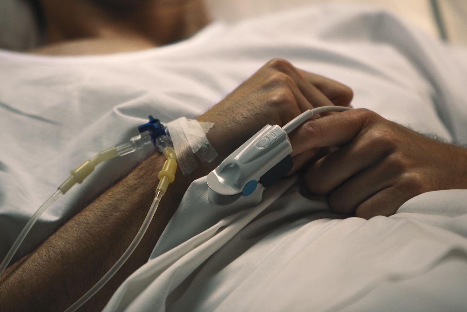 Patients arm with iv line connected to a catheter and tape resting on a hospital bed with hands clasped