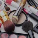 Assortment of makeup products and tools scattered on a white surface including brushes compact powder lipstick nail polish blush pearls and an eyeshadow palette