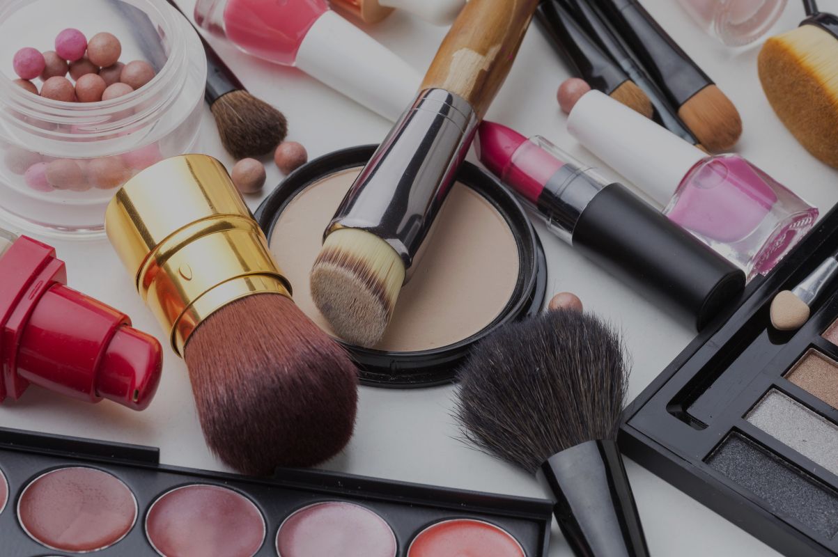 Assortment of makeup products and tools scattered on a white surface including brushes compact powder lipstick nail polish blush pearls and an eyeshadow palette