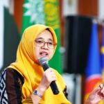 Woman in a yellow hijab and glasses speaks into a handheld microphone on a stage with flags in the background