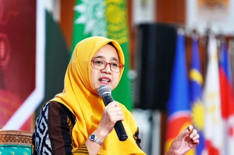 Woman in a yellow hijab and glasses speaks into a handheld microphone on a stage, with flags in the background.