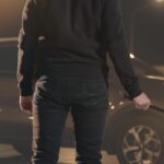 Person in dark clothing seen from the back holding a crowbar beside a car at night