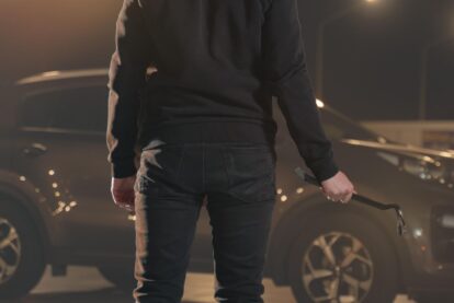 Person in dark clothing, seen from the back, holding a crowbar beside a car at night.