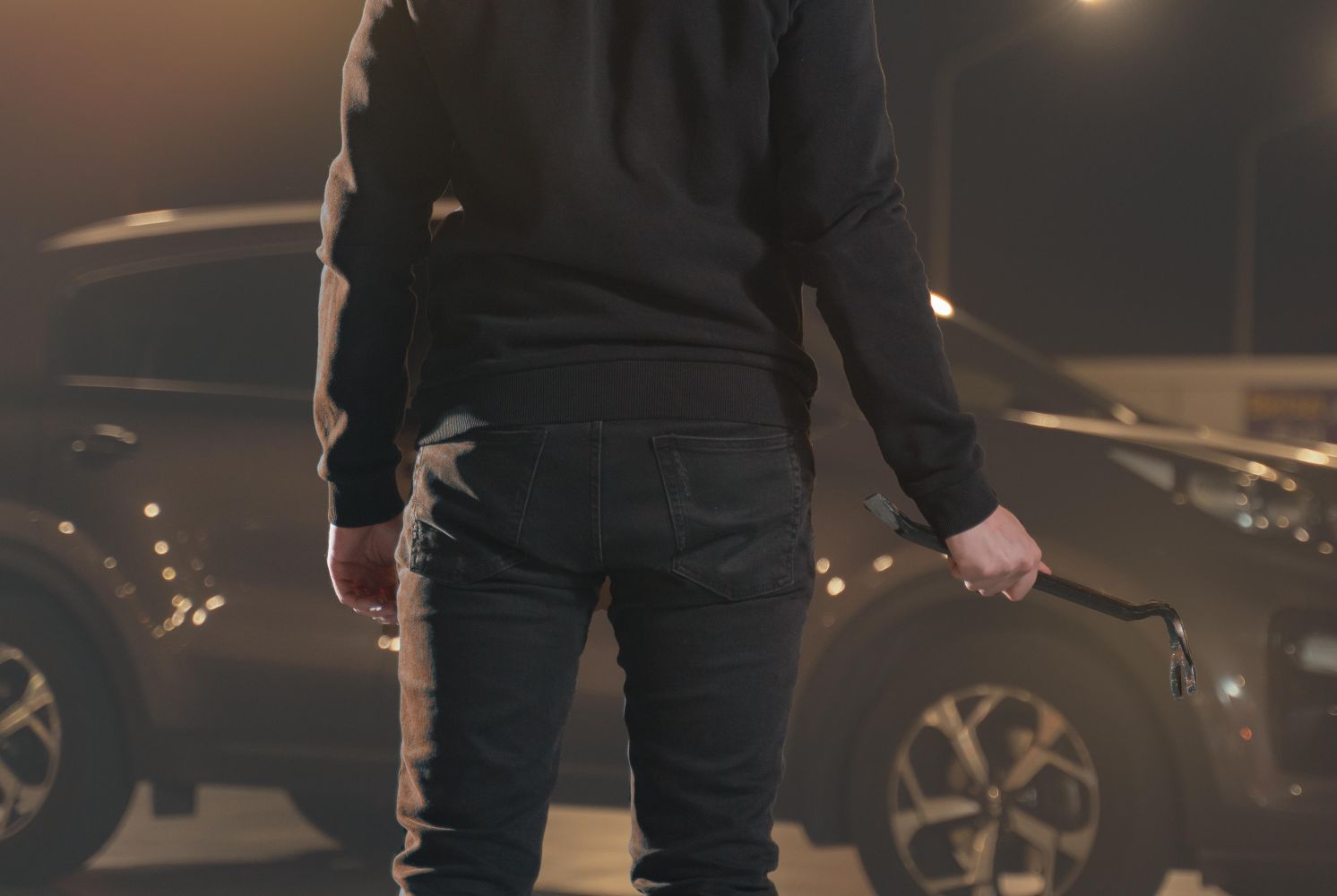 18eklithih catrawarta Person in dark clothing seen from the back holding a crowbar beside a car at night