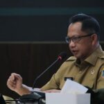 A government official in a tan uniform speaks into a microphone at a panel table reading from papers