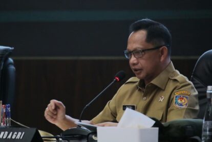A government official in a tan uniform speaks into a microphone at a panel table, reading from papers.