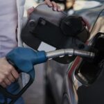 Person holding a blue gasoline nozzle and fueling a car at a gas pump with a white jacket visible in the frame
