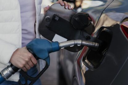 Person holding a blue gasoline nozzle and fueling a car at a gas pump with a white jacket visible in the frame.