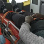 Person lying on a medical stretcher inside an ambulance being attended to by a responder in an orange uniform with blue gloves nearby