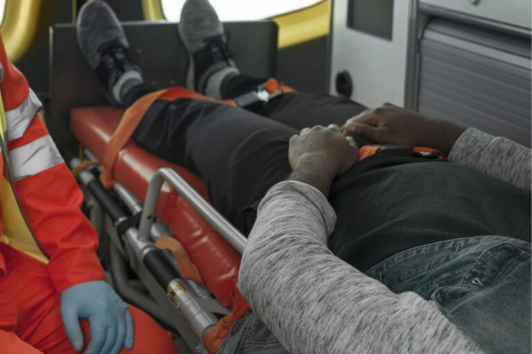 Person lying on a medical stretcher inside an ambulance, being attended to by a responder in an orange uniform with blue gloves nearby.