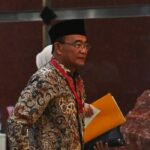 Man wearing a batik shirt and black hat at a conference holding a yellow folder and papers with a red lanyard around his neck