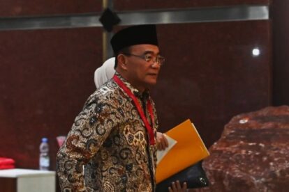 Man wearing a batik shirt and black hat at a conference, holding a yellow folder and papers with a red lanyard around his neck.
