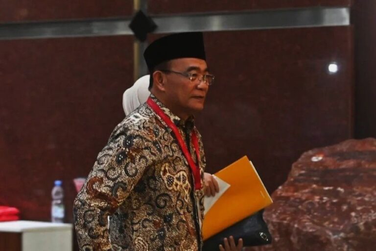 Man wearing a batik shirt and black hat at a conference, holding a yellow folder and papers with a red lanyard around his neck.