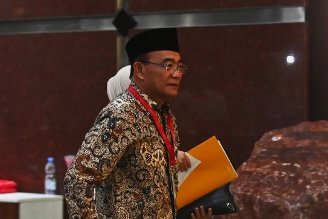Man wearing a batik shirt and black hat at a conference holding a yellow folder and papers with a red lanyard around his neck