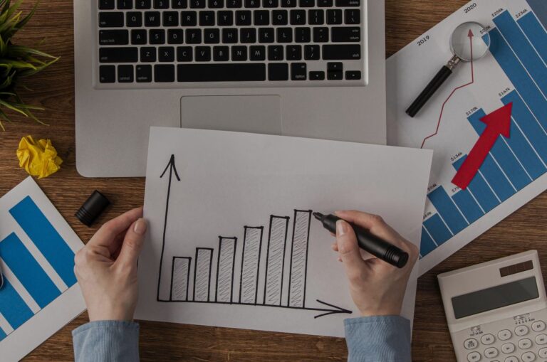 Person draws a rising bar chart on white paper with a marker, on a desk with a laptop and financial charts nearby.