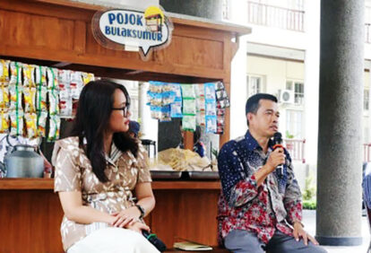 Two people sit by a wooden kiosk outdoors; a man on the right speaks into a microphone while a woman on the left listens.