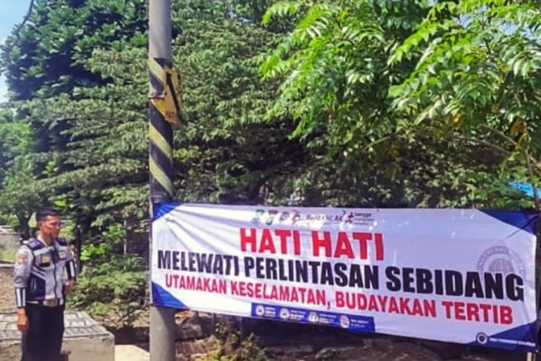 Officer standing beside a safety banner that reads: HATI HATI MELEWATI PERLINTASAN SEBIDANG, UTAMAKAN KESELAMATAN, BUDAYAKAN TERTIB.