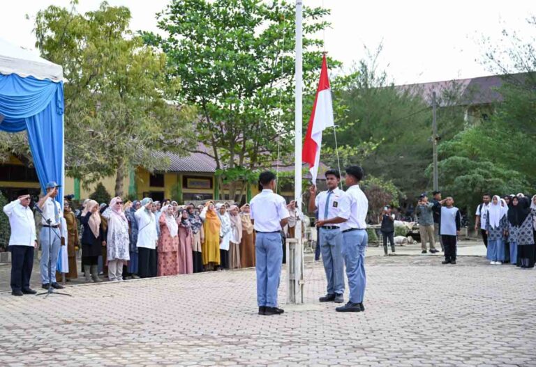 Students in uniform raise and salute as a flag is raised during a school ceremony outdoors while teachers and parents look on.