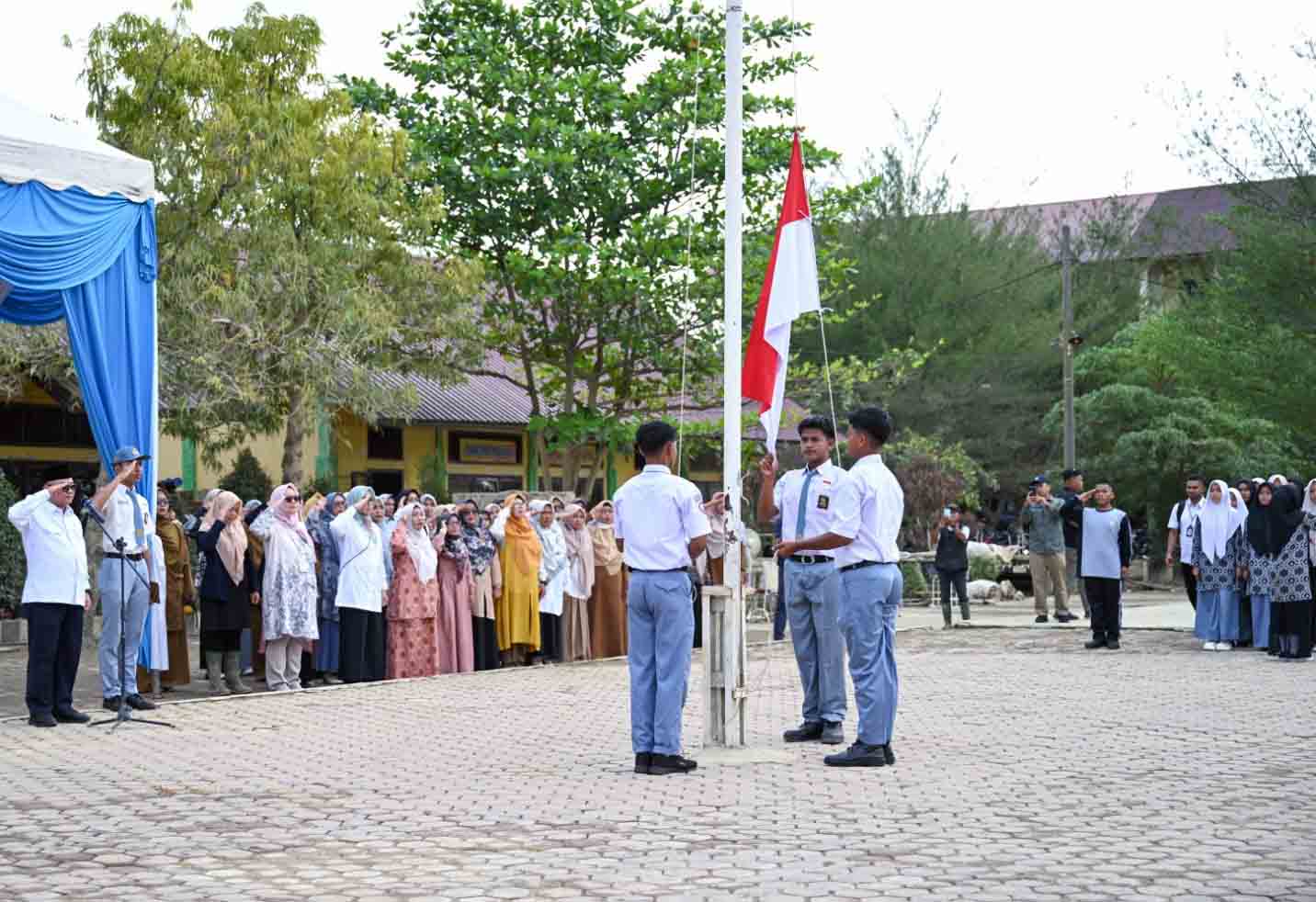 Students in uniform raise and salute as a flag is raised during a school ceremony outdoors while teachers and parents look on