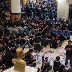 Large crowd of students seated on the floor in a hall listening to a man speaking into a microphone a golden statue sits in the foreground