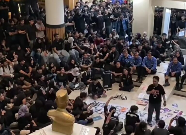 Large crowd of students seated on the floor in a hall, listening to a man speaking into a microphone; a golden statue sits in the foreground.