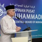 Man in a white embroidered kurta and black cap speaks at a podium with a blue backdrop reading central board of muhammadiah