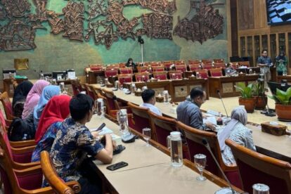 Large conference room with attendees seated around a u shaped table several people wearing hijabs taking notes and using phones in front of glass water pitchers and microphones