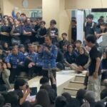A large indoor crowd listening to a speaker holding a microphone with people seated on steps and many standing around some in blue patterned shirts