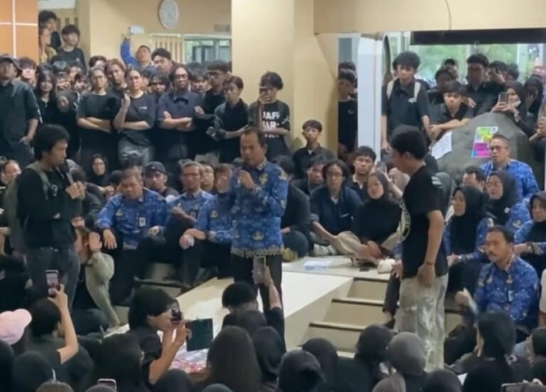 A large indoor crowd listening to a speaker holding a microphone, with people seated on steps and many standing around, some in blue patterned shirts.