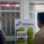 Two men stand near a display of green gas cylinders beside a pertamina sign that reads melayani sepenuh hati