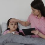 Mother checks her daughters fever in bed placing a hand on her forehead while the daughter lies under a gray blanket and the mother holds a tablet