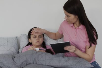 Mother checks her daughter's fever in bed, placing a hand on her forehead while the daughter lies under a gray blanket and the mother holds a tablet.