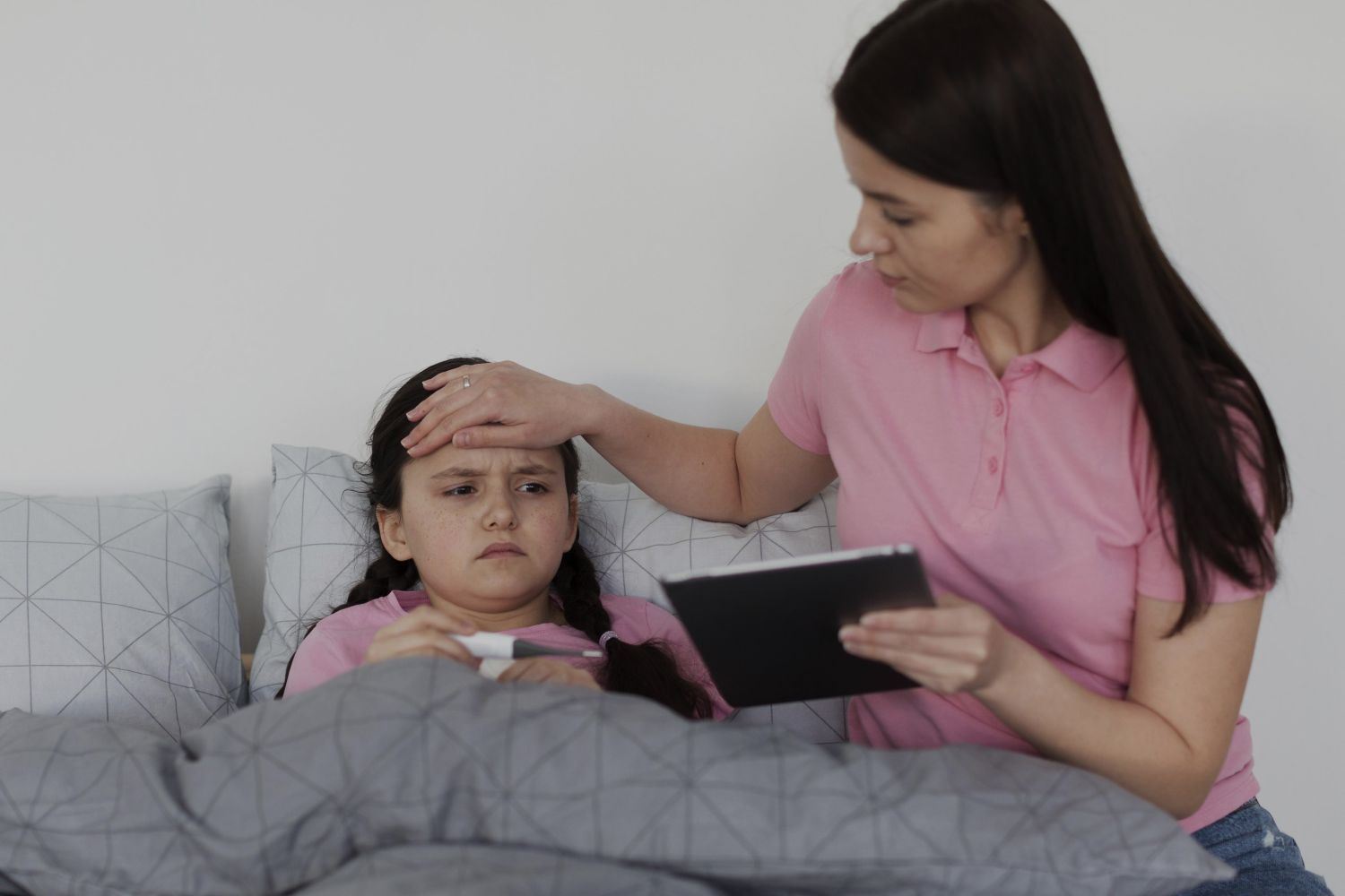 Mother checks her daughters fever in bed placing a hand on her forehead while the daughter lies under a gray blanket and the mother holds a tablet