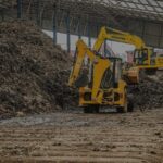 Two yellow excavators work on a large pile of wood waste inside a metal framed warehouse dirt floor in the foreground