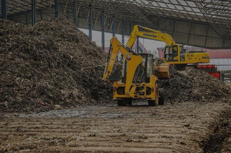 Two yellow excavators work on a large pile of wood waste inside a metal framed warehouse dirt floor in the foreground