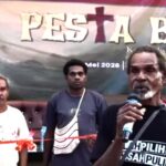 Man speaking into a microphone on a stage flanked by two men with a banner in the background showing event text