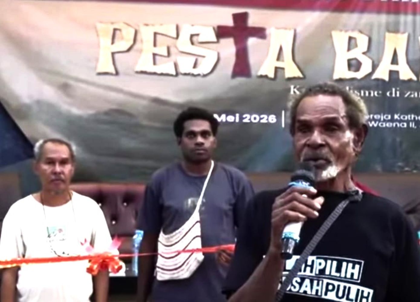 Man speaking into a microphone on a stage flanked by two men with a banner in the background showing event text